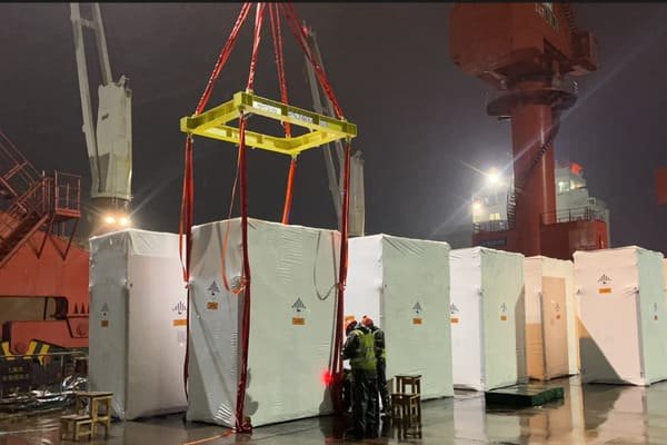 A large, container-sized energy storage system being lifted by a crane at a port.